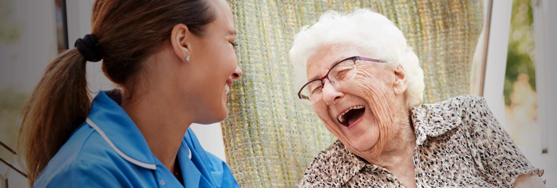 A senior woman and a young caregiver laugh together.